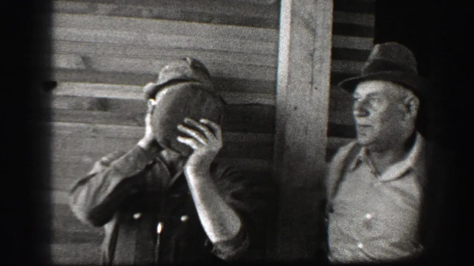 1939 California home-movie frame of a man holding a jug to his face near wood siding while another adult man in a dark hat stands beside him.
