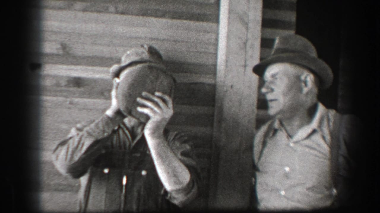 Two men in hats stand by a wood building as one drinks from a large jug.