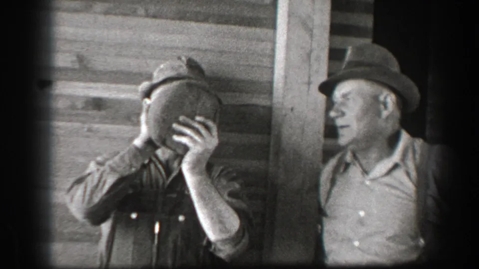 In 1939 California home-movie footage, two adult men in work clothes stand outside a wood building while one raises a jug to his mouth.