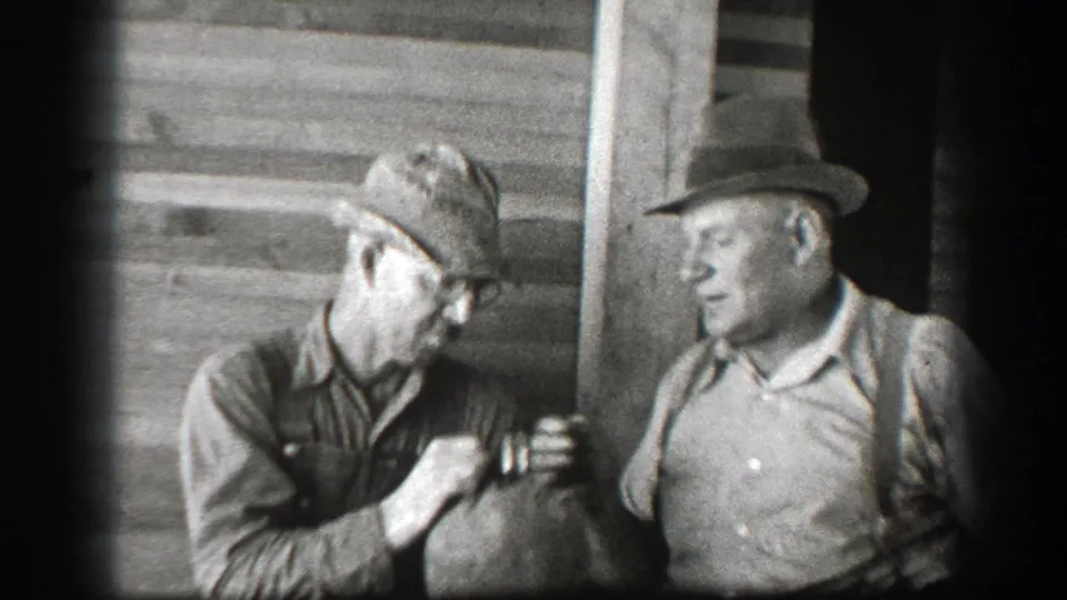 Two men in hats stand near wooden siding in 1939 California footage as the man holding the jug turns toward his companion.