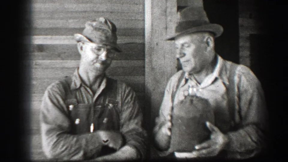 The men pause together outside the wooden building, with one holding the jug and the other looking on in work clothes and a cap.