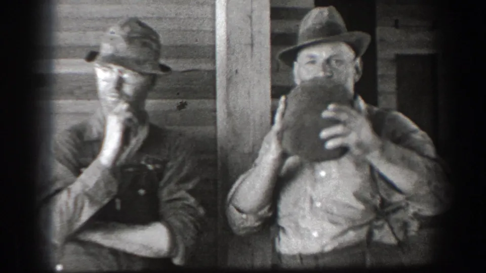 Two adult men in period workwear stand beside a wooden structure in California home-movie footage, one lifting a jug to drink.