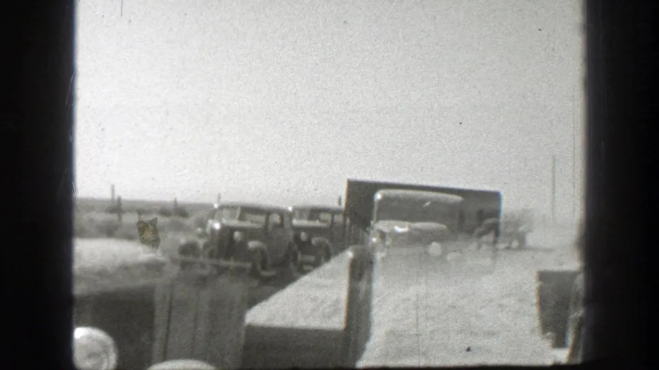 From near a truck hood, the home-movie camera looks toward other vintage trucks moving through dust on a California dirt road.