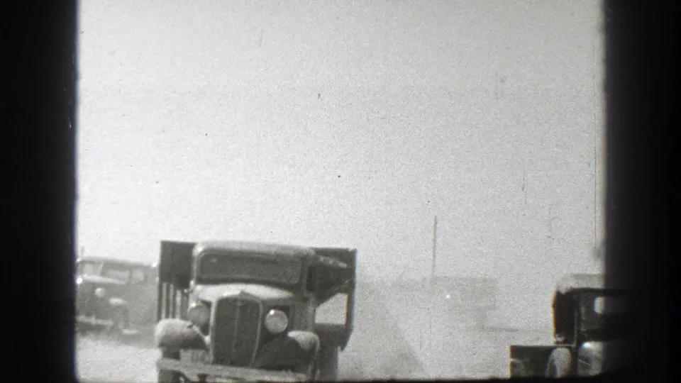A vintage truck faces the camera on a dusty California road, with another truck partly visible behind it in the haze.