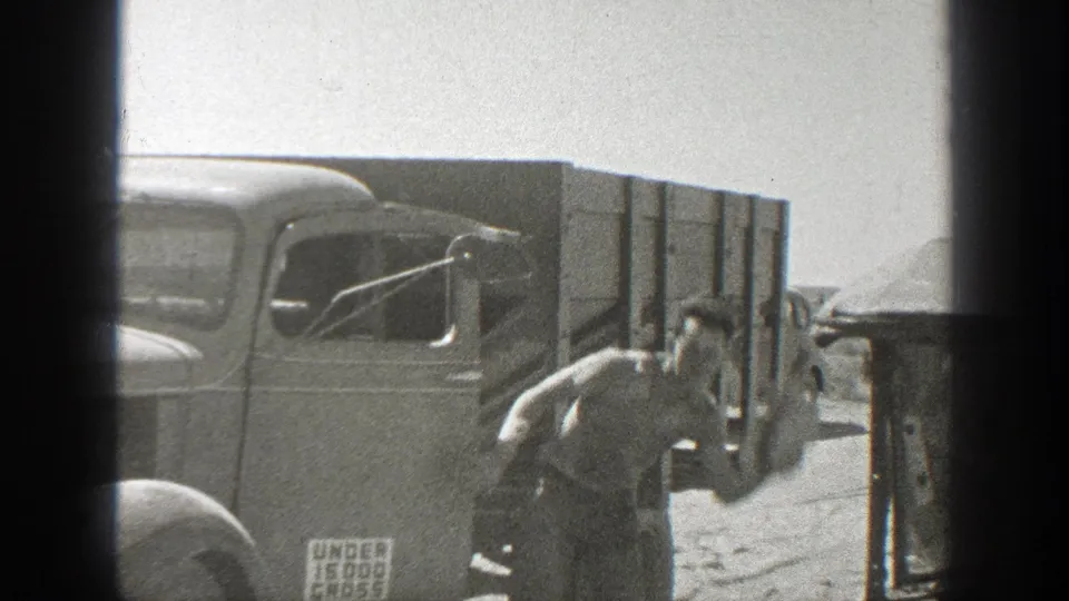 A man in a brimmed hat stands or steps near an older truck on a dusty California road in 1939 footage.