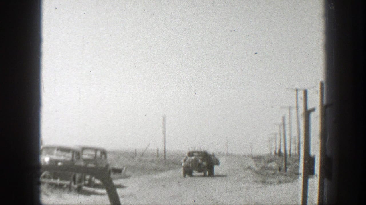Dusty dirt road with parked vintage cars, a distant truck, and roadside utility poles.