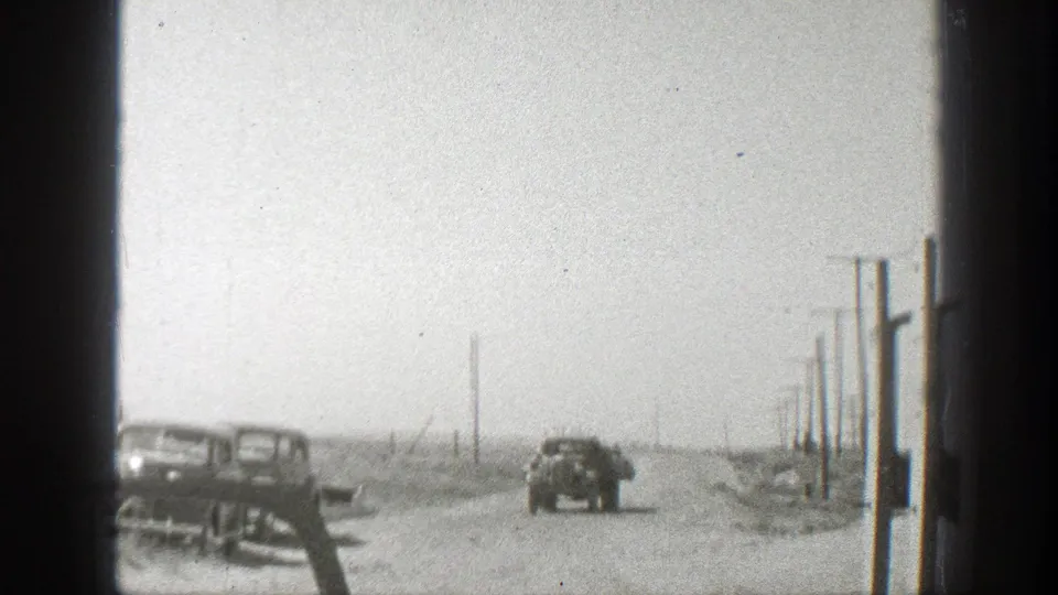 1939 California home-movie frame showing a dusty road, parked automobiles, a truck driving away, and utility poles in an arid landscape.