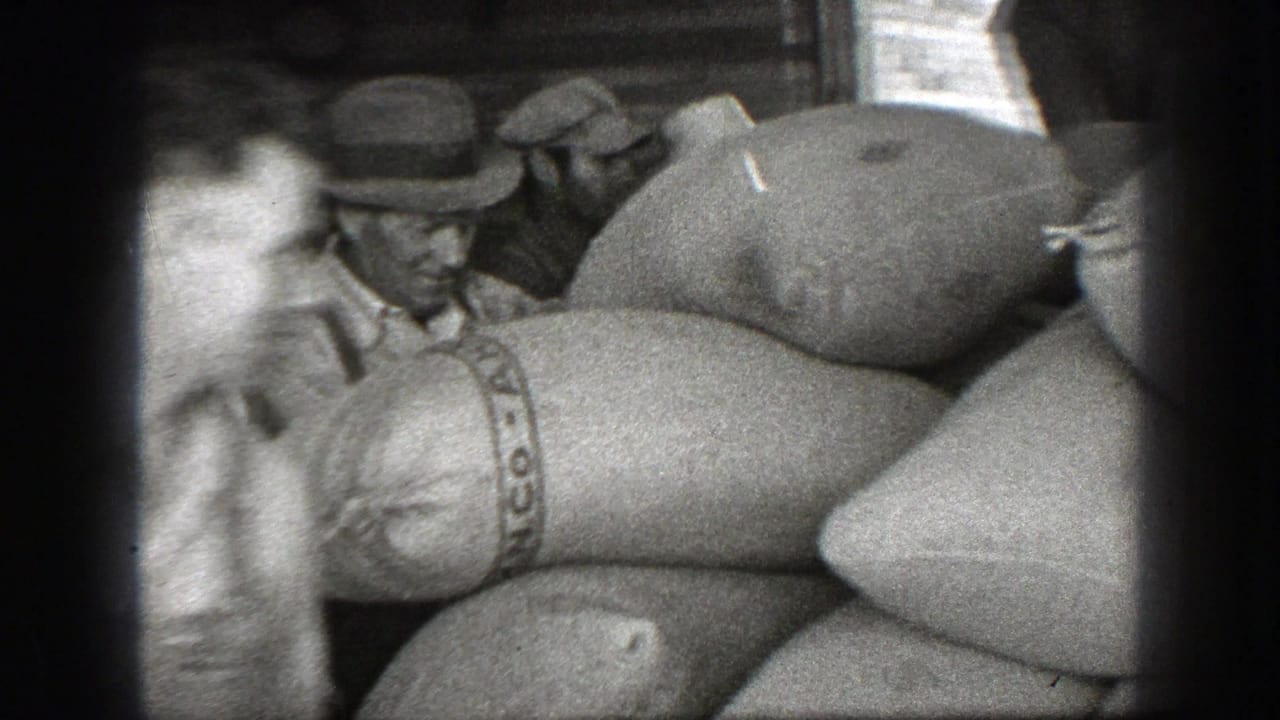 Men in hats stand beside stacked cloth sacks inside a wooden building.