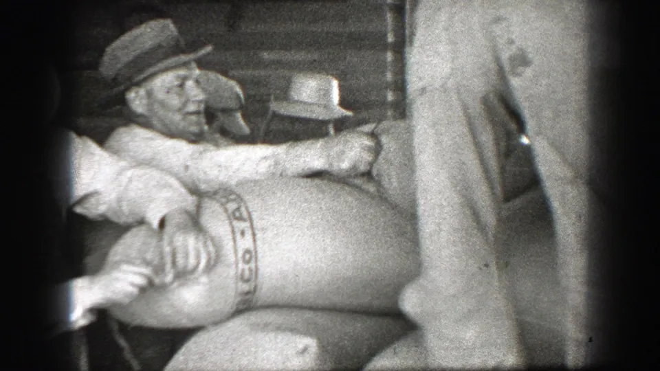 Men in work clothes handle a large bag marked CO-OP, with a standing figure partially blocking the close warehouse view.
