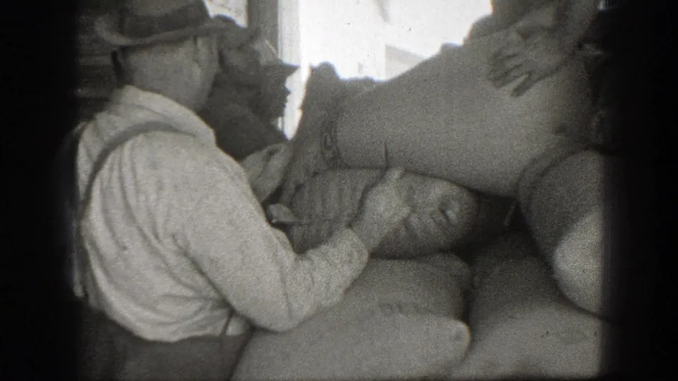 1939 California home-movie frame of a worker in a hat and suspenders standing beside large burlap-like sacks.
