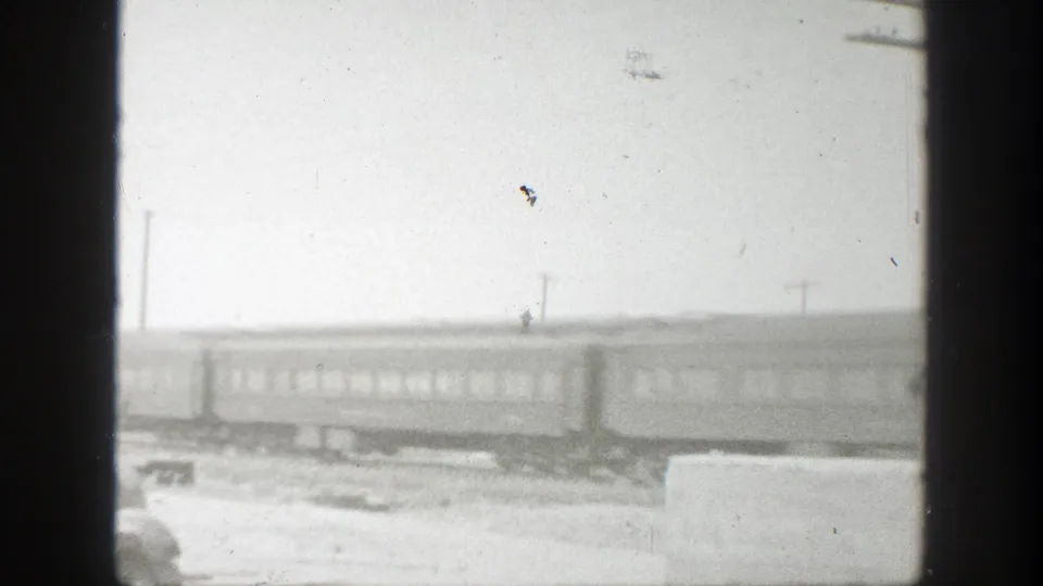 Black-and-white home-movie frame of passenger rail cars passing through rural California terrain under a pale, overcast sky.