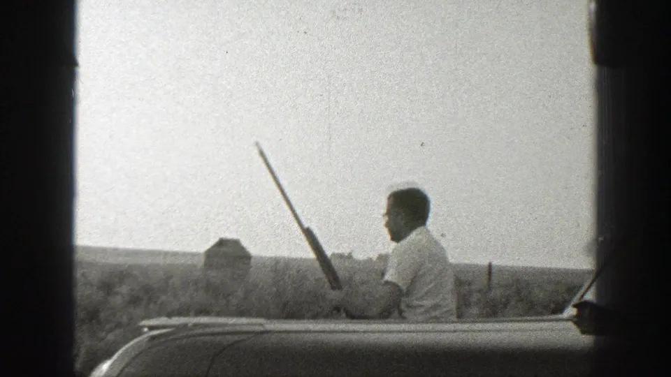 1939 California home-movie frame showing a man in profile holding a long gun beside a parked automobile in an open rural setting.