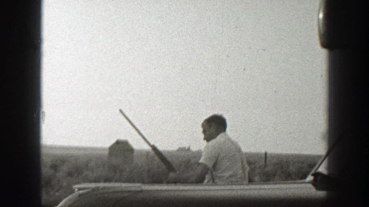 Man in a light shirt holds a long gun beside open fields, seen from inside or beside a vehicle.