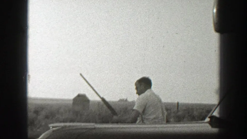 In 1939 California home-movie footage, an adult man with a long gun stands near open rural fields with small structures in the distance.