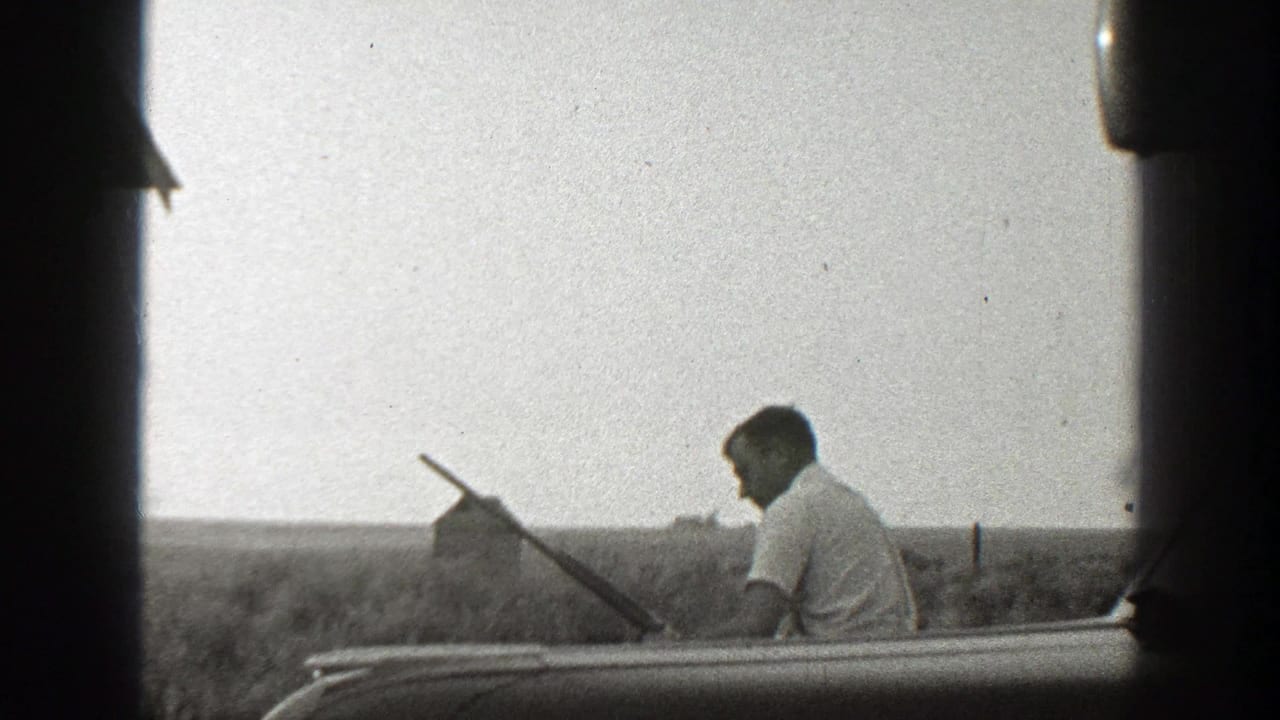 Man with a long gun walks along a rural field with a small barn-like structure in the distance.