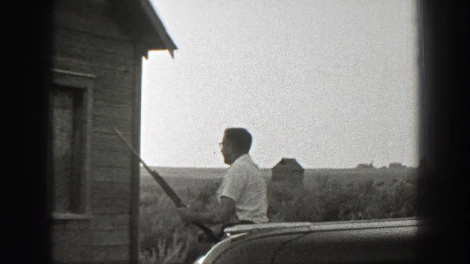 A 1939 California home-movie frame shows a man with a long gun near a wooden rural building and open fields beyond.