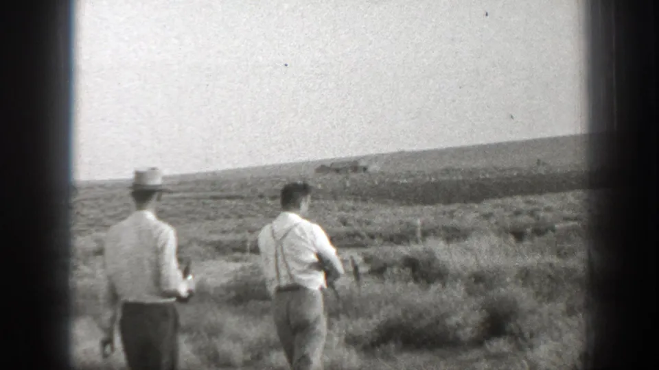 Two adult men in period clothing walk away across an open California field, with one man visibly carrying a rifle.