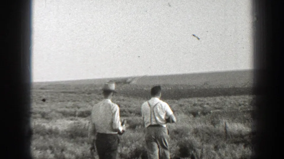 Two men in 1939 California rural footage continue across a scrub field, framed by dark 8mm film edges and distant hills.