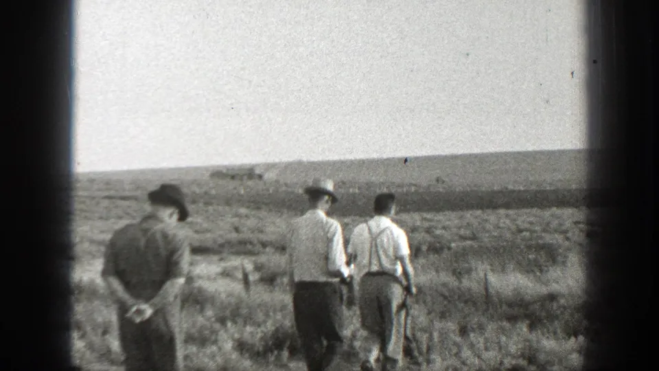 Three adult men cross an open rural California field, including one darker-clothed man trailing with his hands behind his back.