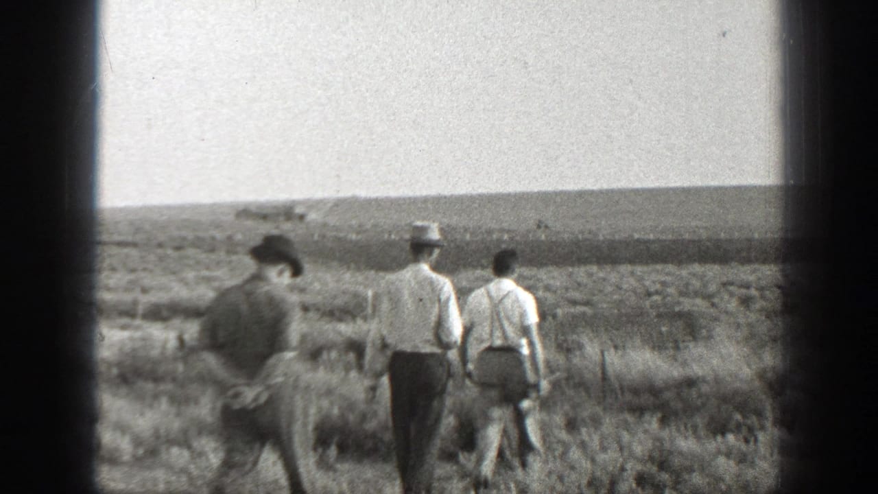 Three men walk away across a dry open field, two wearing hats and one in suspenders.
