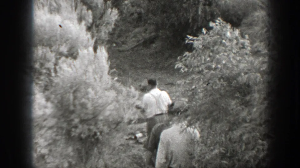 Men continue along a shaded brush-lined path, with soft grain and handheld framing typical of 8mm home movies.