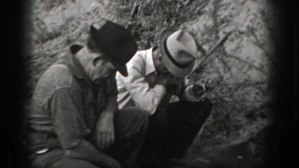 Three men in period outdoor clothing crouch in a wooded area, appearing to examine something on the ground together.