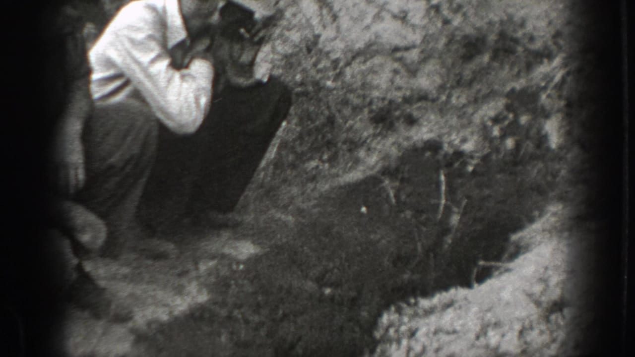 Man in suspenders crouches or steps near a rocky wooded bank in black-and-white footage.