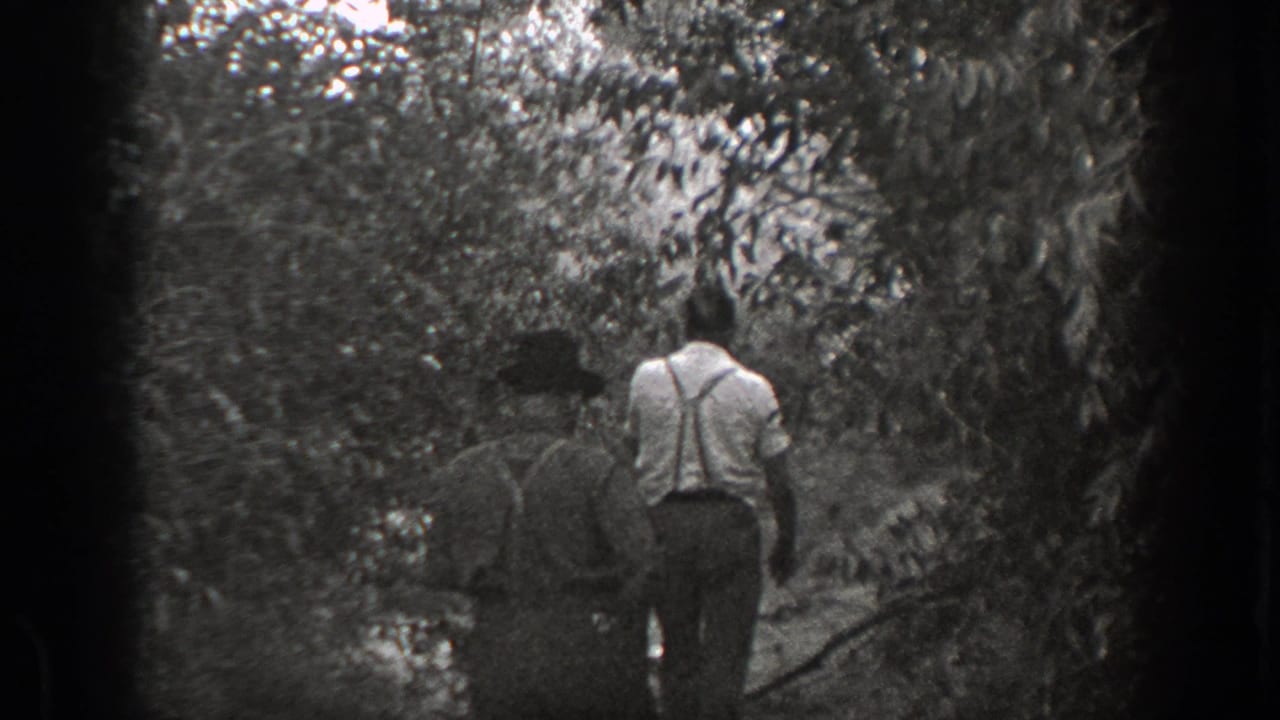 Two men walk away along a narrow path through dense trees and brush.
