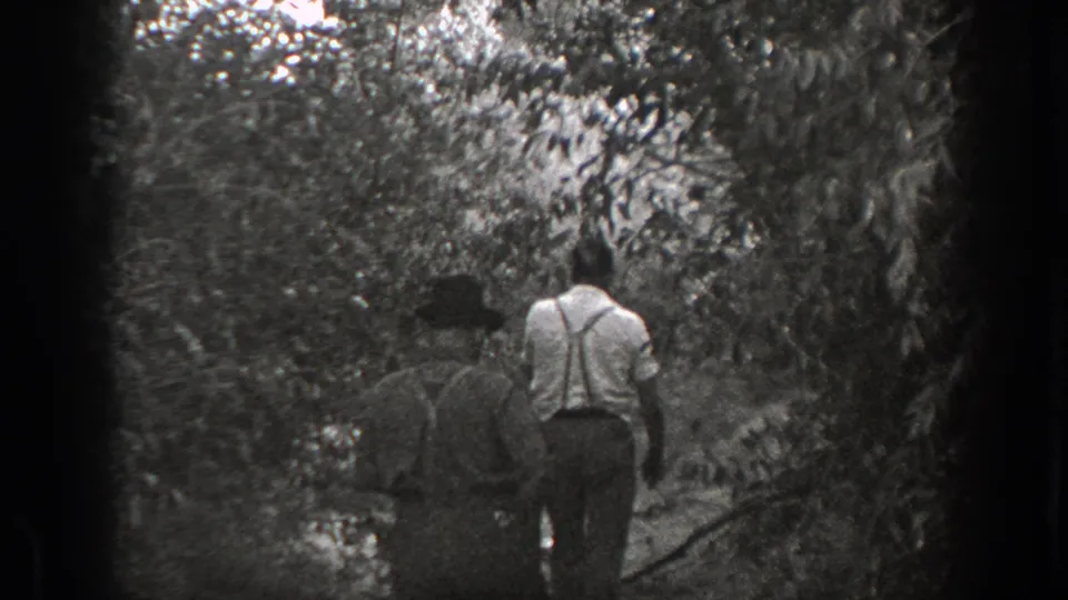 1939 California home-movie frame with two men, one in a hat and one in suspenders, walking away through dense woodland.