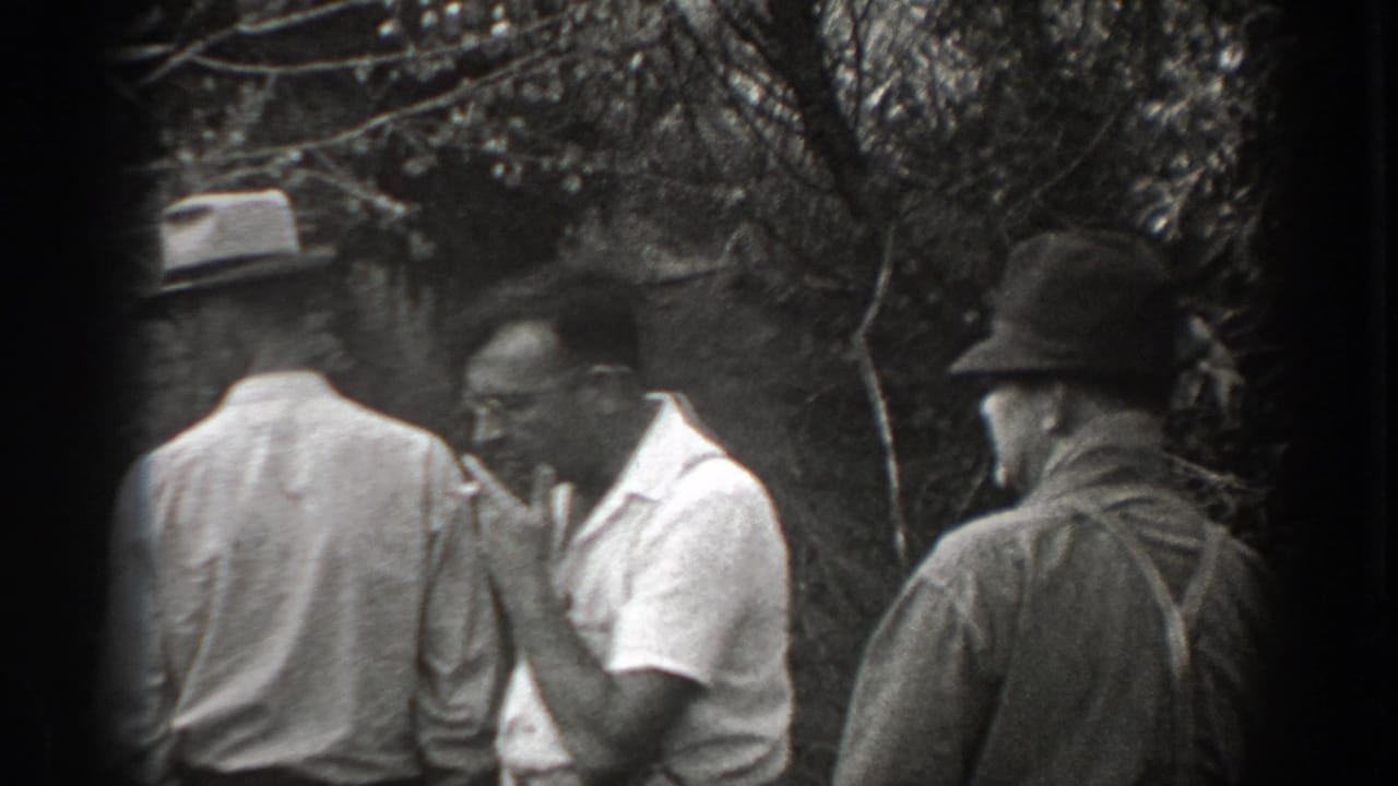 Men standing near bushes, with one man smoking in a light shirt.