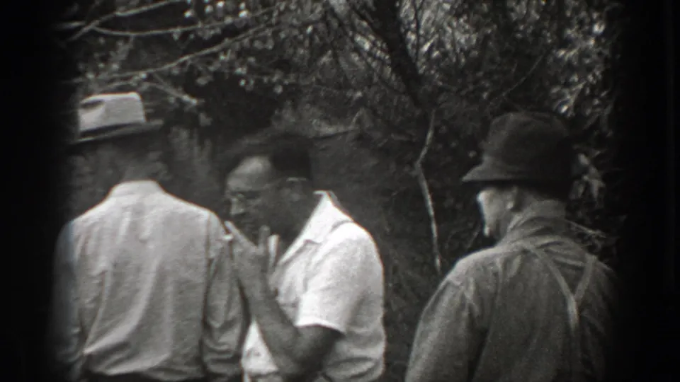 Adult men stand close together near dense brush in California home-movie footage, including a man in a light shirt holding a cigarette near his mouth.