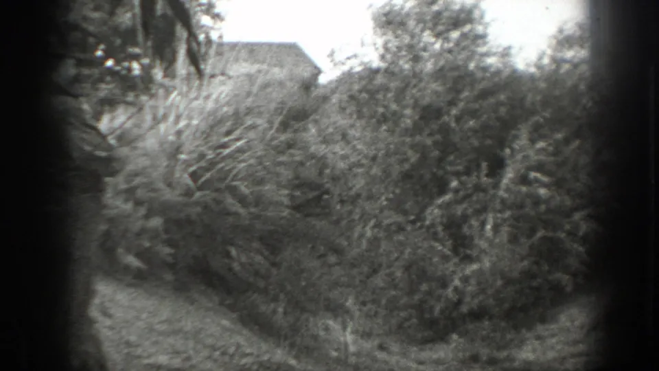 A narrow outdoor path curves through thick bushes, with adult men partly visible at the left edge and a building roofline faintly seen beyond the vegetation.