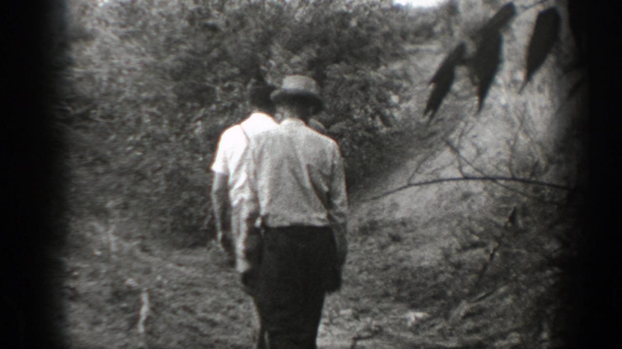 Two men walk away on a narrow path with brush and a hanging branch nearby.