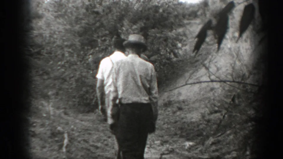 Two men in light shirts continue along a California rural path, with foliage and a dark branch at the edge of frame.