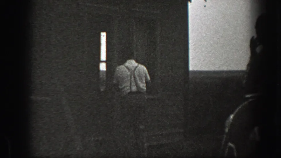 Inside the dim wooden shack, a man in a light shirt and suspenders faces a dark doorway in 1939 California footage.