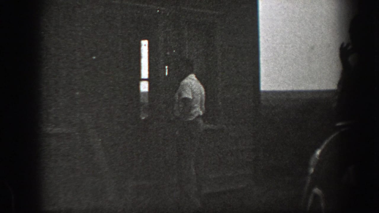 Grainy 1939 California 8mm frame showing a man in a light shirt near house steps, partly obscured by a dark foreground edge.