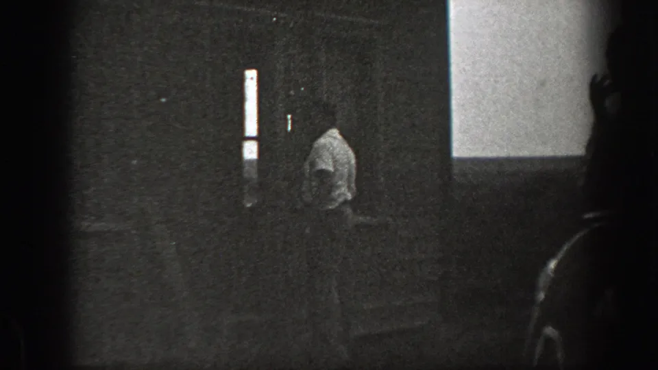 Grainy 1939 California 8mm frame showing a man in a light shirt near house steps, partly obscured by a dark foreground edge.