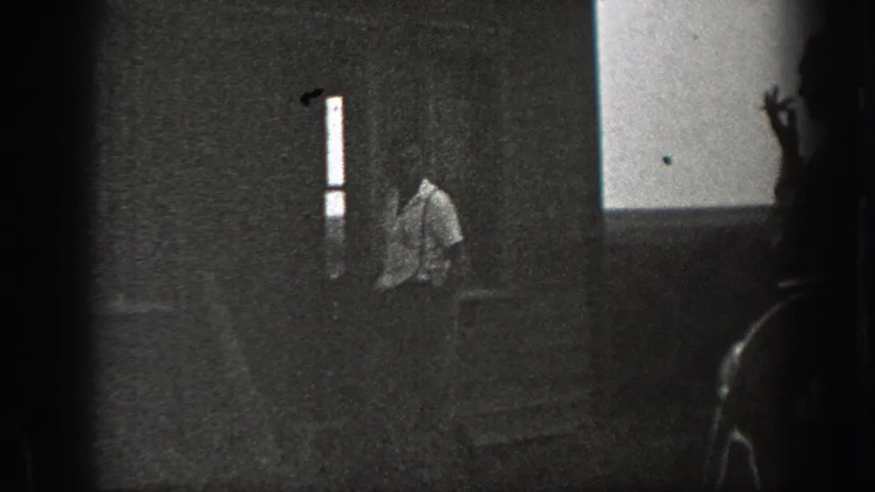 1939 California home-movie frame with a man near house steps, viewed through a dark foreground shape and a bright vertical opening.