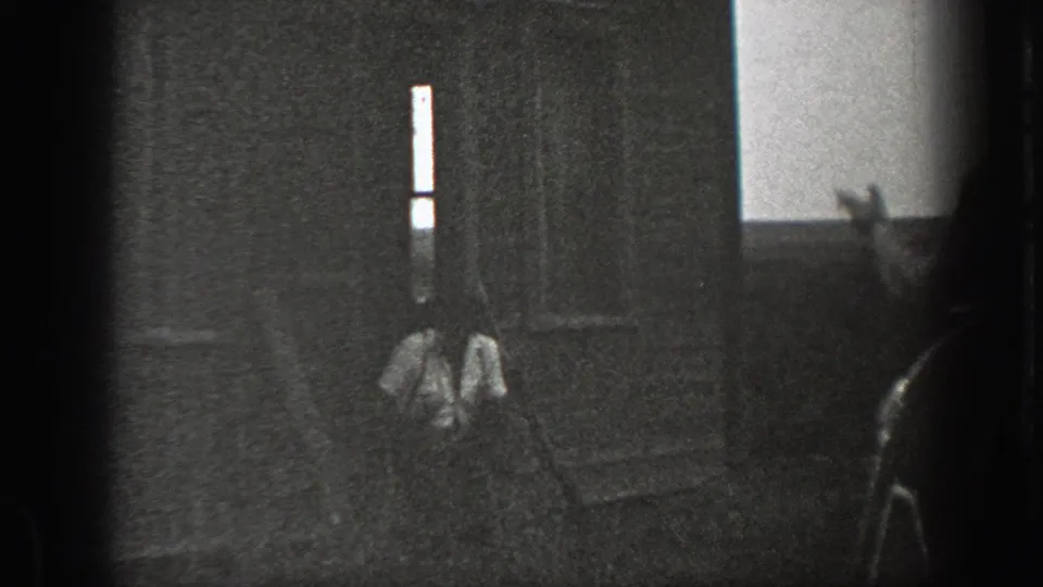 Grainy 1939 California 8mm frame showing a man seated on house steps with a raised hand visible in the foreground.