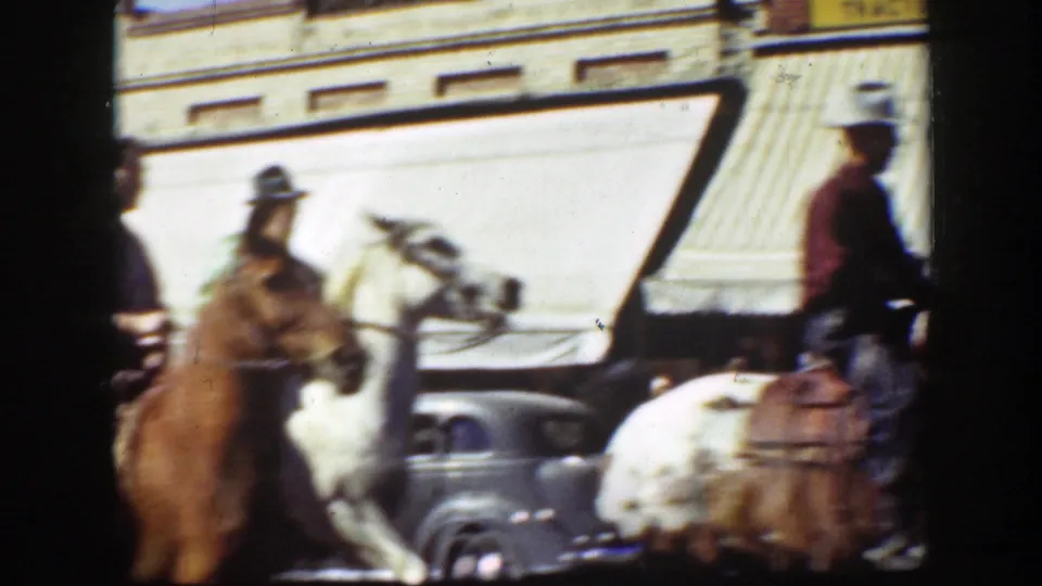 Color 8mm frame from California, 1939, showing horseback riders in hats near parked period automobiles and a shop awning.