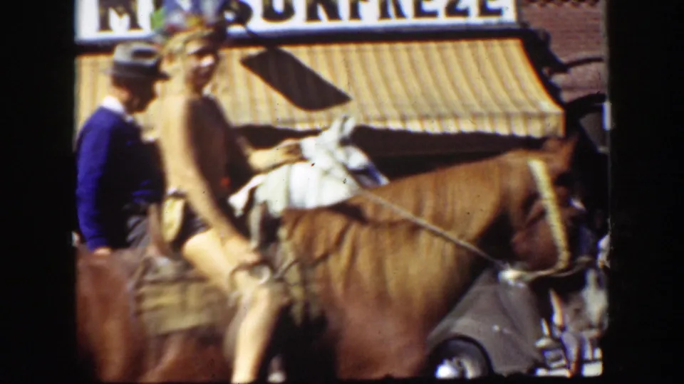 1939 California parade view of riders on brown horses passing a storefront awning, with faces and commercial signage partly visible.