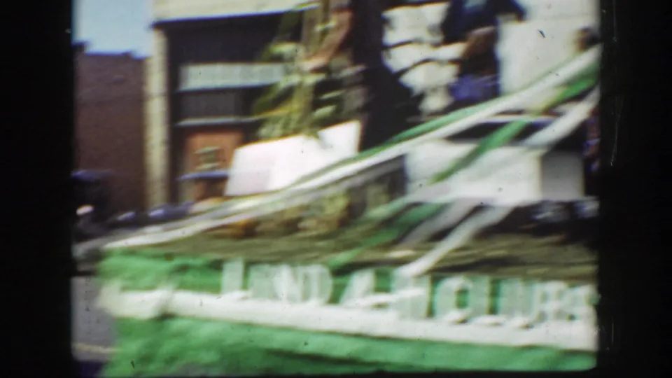 Motion-blurred 1939 California parade frame with a green structure or banner moving past storefronts and a partial figure.