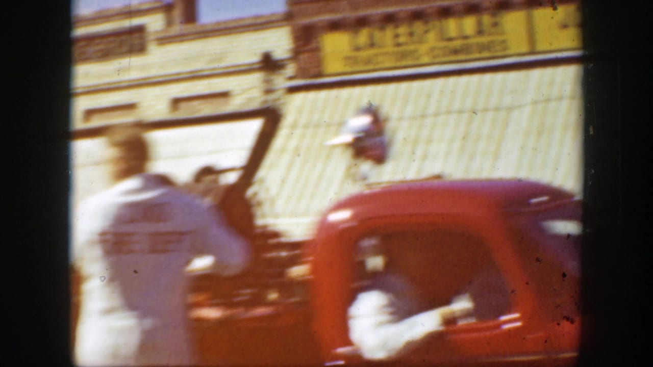 1939 California street view with a red truck, storefront awning, yellow implement sign, and pedestrian movement in soft 8mm focus.
