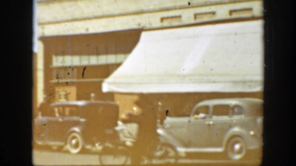 California street scene with late-1930s automobiles, storefront awnings, and pedestrians visible through faded 8mm film.