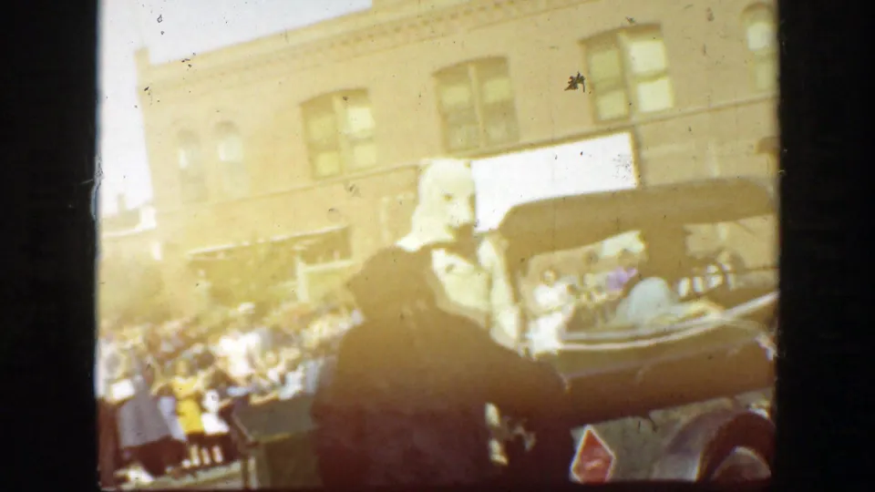 1939 California crowd scene beside a parked vehicle, with a light-haired or costumed figure in the foreground and storefronts behind.