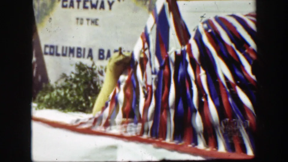 1939 California home-movie frame showing red, white, and blue bunting on a float with a visible Gateway to the Columbia Basin sign behind it.
