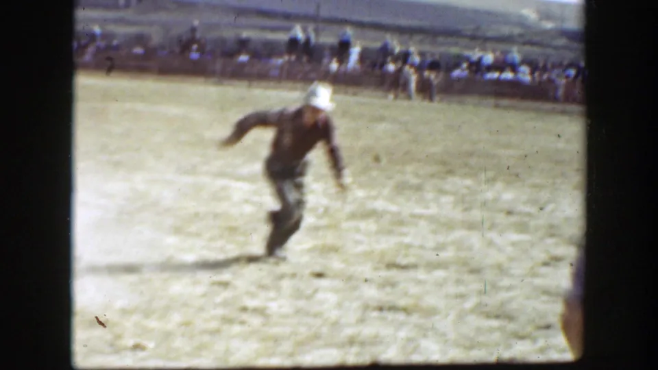 1939 California rodeo frame showing a cowboy in a white hat moving away across the dirt arena, with spectators beyond the rail.