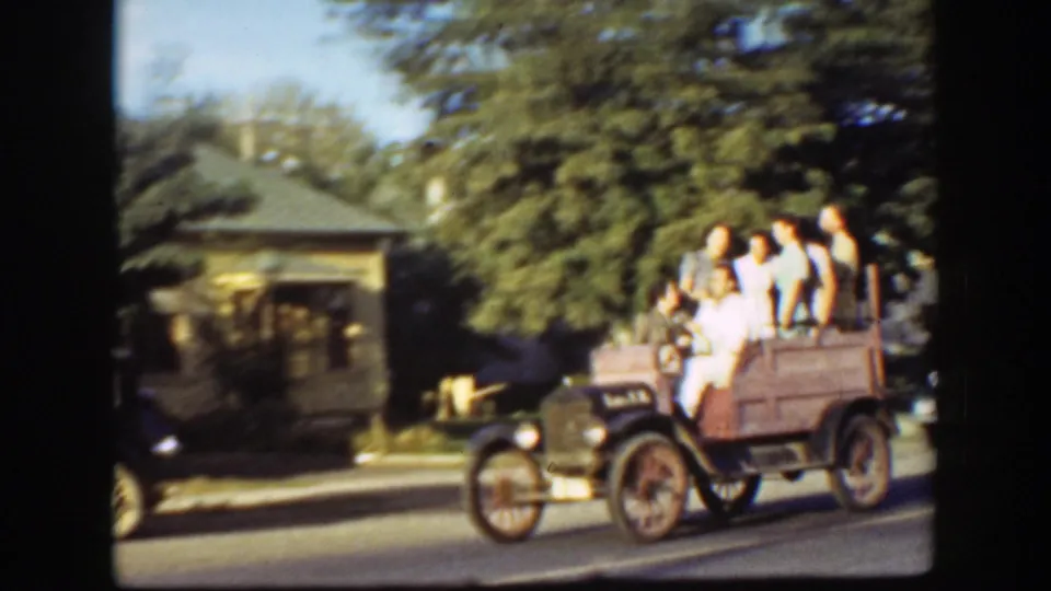 1939 California home-movie frame showing several passengers riding in an open vintage automobile past trees and a roadside building.