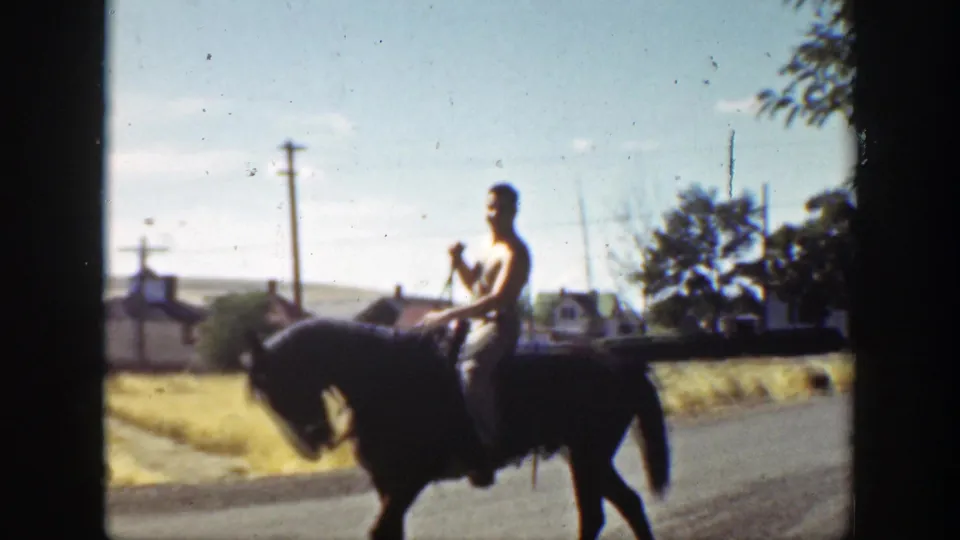 A rider on a dark horse moves along a California roadside, with small houses, utility poles, and open land visible.