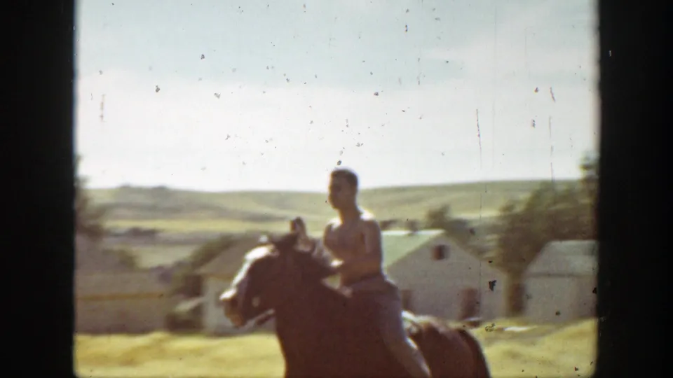 Shirtless rider on a brown horse moves through a sunlit California field with low hills and trees in the background.